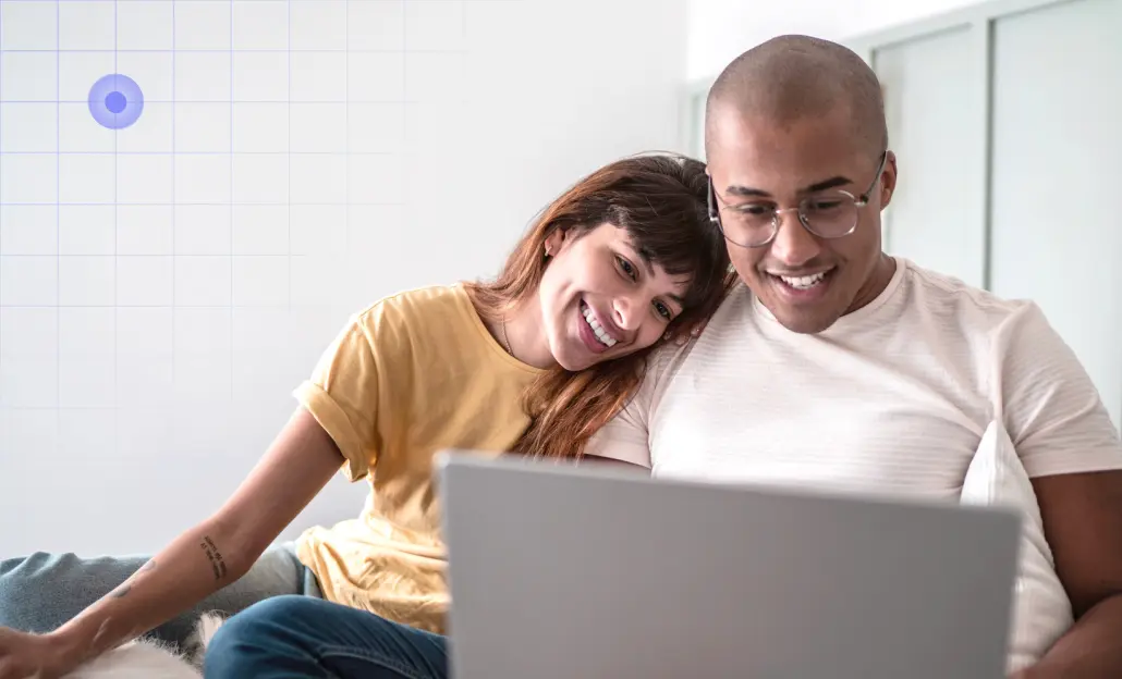 A smiling woman leans on a man's shoulder as they look at a laptop together in a brightly lit room.